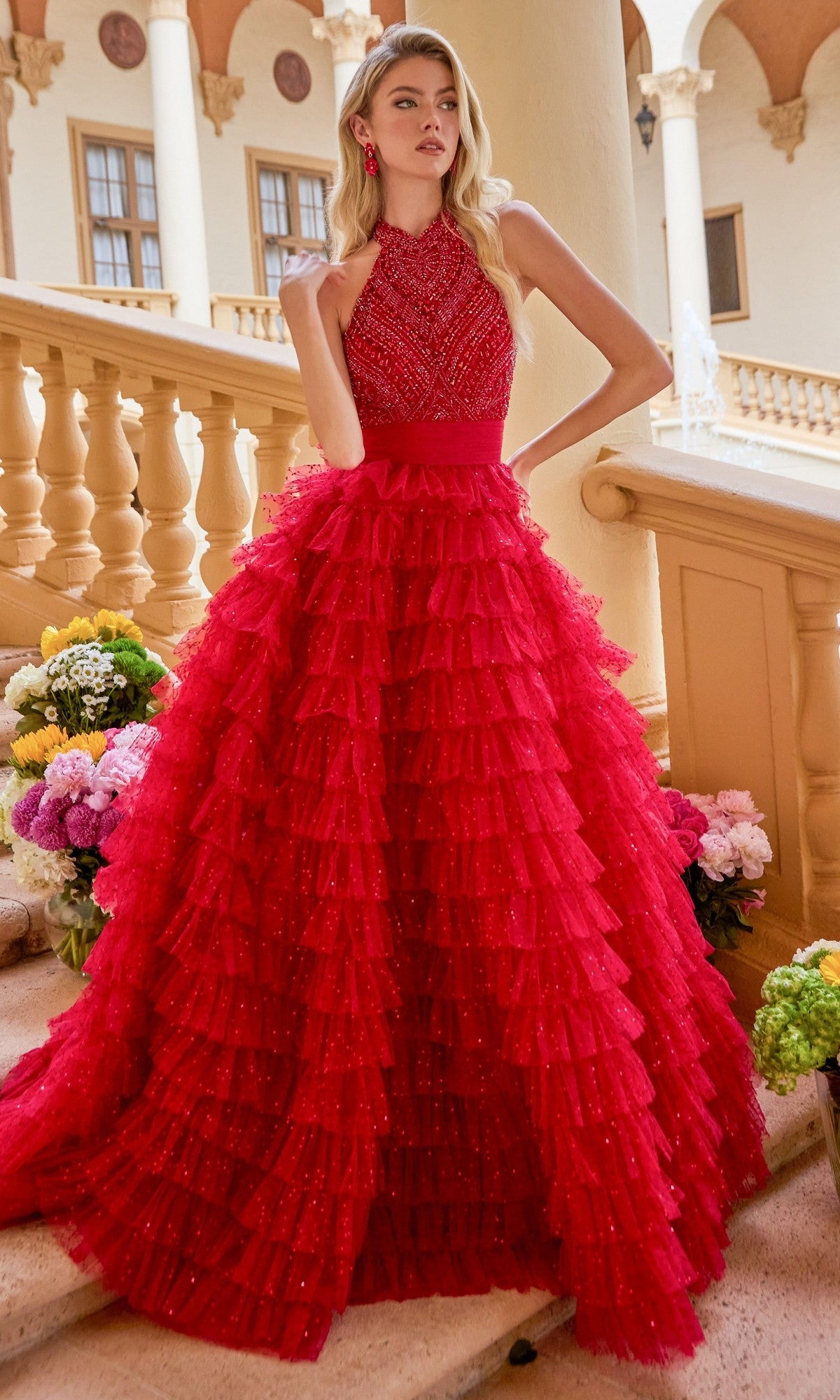 A woman stands on stairs surrounded by flowers, wearing the Ava Presley 47258 long prom dress featuring a sleeveless, beaded halter bodice and a red tiered ruffled ball gown skirt.